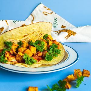 Een close-up van een open mais taco met daarin zoete aardappel blokjes, harissa tofugehakt en krulpeterselie op een blauwe achtergrond met een theedoek er achterlangs gedrapeerd