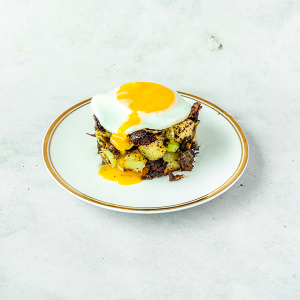 Een wit bord met een gouden randje met daarop Boudin Noir hash met spruitjes en een gebakken ei met een zachte dooier op een wit marmeren achtergrond.