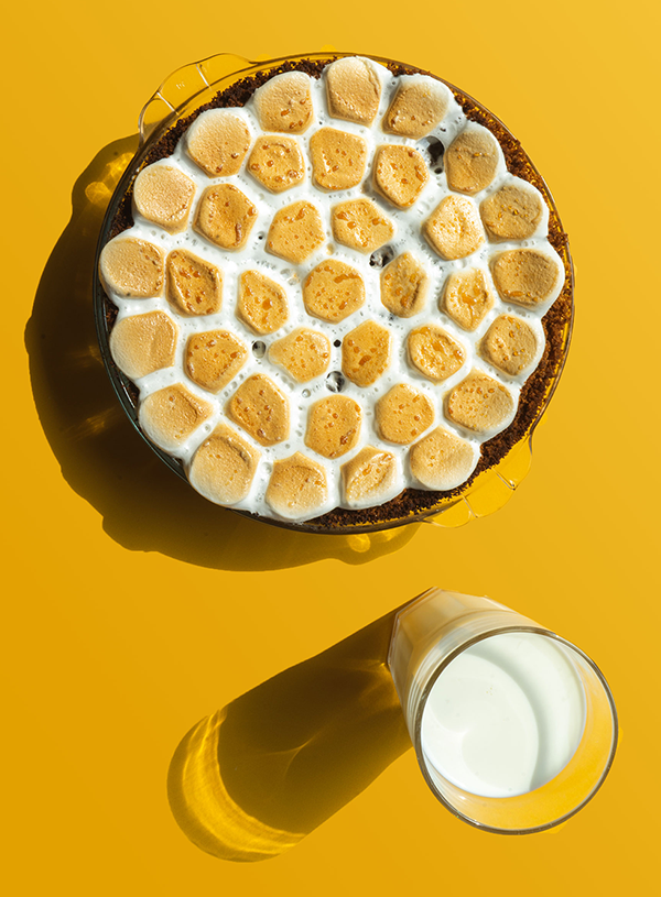 Een foto van bovenaf genomen van een smores pie bedekt met gebrande marshmallows met een glas melk ernaast op een gele achtergrond.