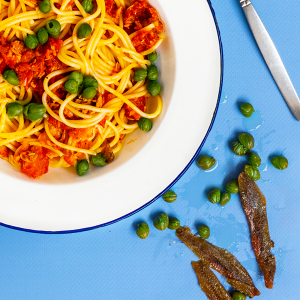 Een close-up van een wit bord met spaghetti met rode saus en groene kappertjes op op een blauwe achtergrond met ernaast een vork, een open blik tomaten, wat losse kappertjes en ansjovisfilets en het woord PUTA gespeld in spaghetti.