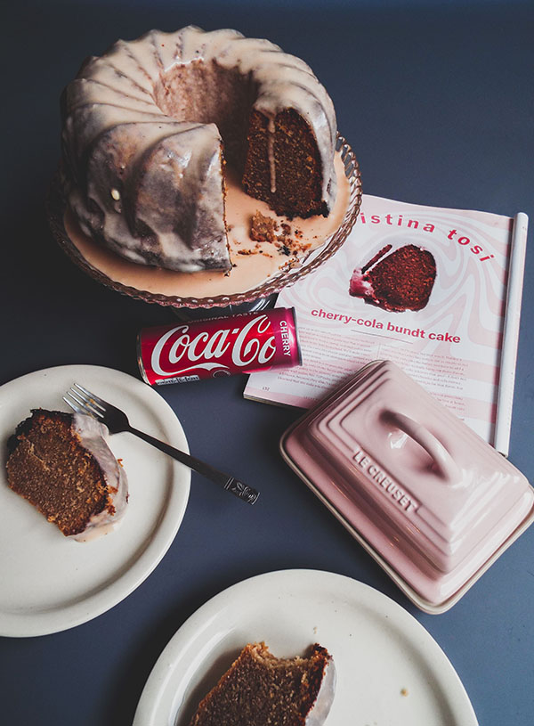 Cherry cola cake op een cake standaard, met erbij twee bordjes met plakjes van de cake, een roze botervloot en een tijdschrift opengeslagen op het recept.
