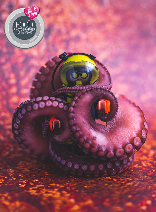 Een Playmobil duiker omringt door gekookte rode tentakels van een octopus op een rood gemêleerde achtergrond.