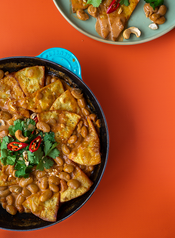 Een grote pan met lima bonen penang chilaquiles, met daarop wat koriander en boven in beeld nog een bord met hetzelfde gerecht erop. Op een oranje achtergrond.